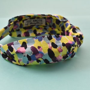 Puede incluir: Una diadema colorida con un diseño anudado. La tela presenta un patrón vibrante de pinceladas amarillas, negras, moradas, azules y rosas. El texto "TOT KNOTS of Brighton" es visible en una pequeña etiqueta dentro de la diadema.