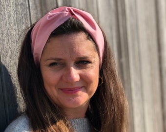 Puede incluir: Una mujer con el pelo largo y castaño lleva una diadema de satén rosa claro. La diadema está anudada en la parte superior. Lleva un jersey gris. El fondo es una valla de madera.