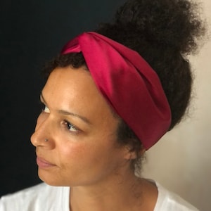 Op de afbeelding: Een bordeauxrode satijnen hoofdband wordt over het voorhoofd gedragen. De hoofdband is aan de bovenkant gedraaid en bedekt het haar. De persoon draagt een wit T-shirt.