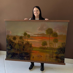 May include: A landscape painting depicting a sunset over a swamp with palm trees. The artwork features a warm color palette of orange, yellow, and green. The painting is framed with a dark wooden border.