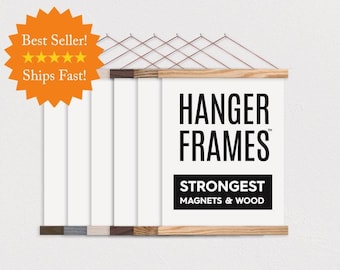 May include: A set of wooden poster hanger frames with magnetic closures, featuring a white display sheet printed with the black text HANGER FRAMES STRONGEST MAGNETS & WOOD, suspended by thin copper-colored wires against a plain white background.