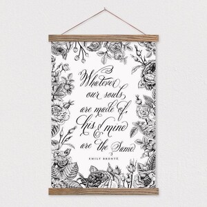 May include: A black and white floral print wall hanging with the quote "Whatever our souls are made of, his & mine are the same" by Emily Bronte. The print is framed by a floral border and held by a wooden hanger.