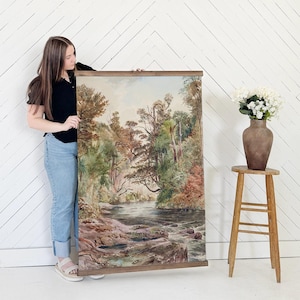 May include: A large landscape painting depicting a river flowing through a forest. The artwork features a variety of trees with green, brown, and red foliage. The painting is framed with a wooden border and is being held up.