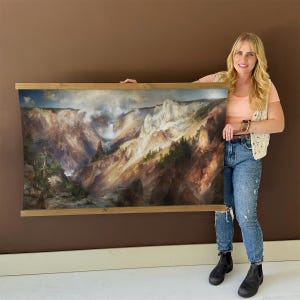 May include: A landscape print of a canyon with a waterfall, framed by a light brown wooden frame. The artwork depicts a detailed view of a canyon with a waterfall, trees, and a cloudy sky. The print is held up by a person.