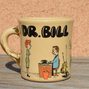 May include: A beige ceramic mug with a black handle and a cartoon illustration of a man and woman. The mug has the text "DR. BILL" printed in black on the front.