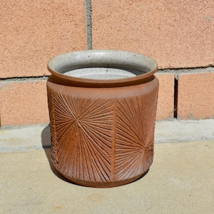 Op de afbeelding: Cilindrische keramische pot in terracotta kleur met een getextureerd, gesneden ontwerp. Het ontwerp heeft stralende lijnen die uitgaan van een centraal punt, waardoor een patroon ontstaat. De rand is een lichtere, gespikkelde grijze kleur.