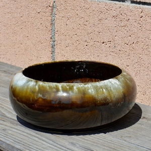 Puede incluir: Un cuenco de cerámica vidriada, de forma ancha y redondeada. Presenta un acabado brillante con tonos marrones, verdes y blancos. Se encuentra sobre una superficie de madera envejecida, con una pared texturizada al fondo.