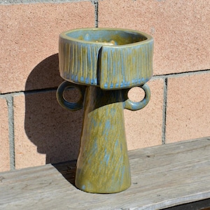 May include: A ceramic vase with a wide, cylindrical top section featuring vertical etched lines and a tapered base. The piece is finished in a mottled glaze of olive green and dusty blue, and includes two small, circular handles on the sides.