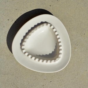 May include: White ceramic ashtray with a triangular shaped depression in the centre. The ashtray has a scalloped edge.