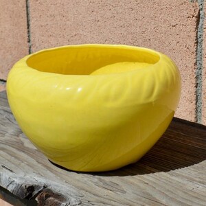 May include: A bright yellow ceramic bowl with a smooth, glossy finish and a rounded, bulbous shape. The exterior features a subtle, embossed leaf-like pattern that wraps around the circumference of the vessel, creating a textured appearance.