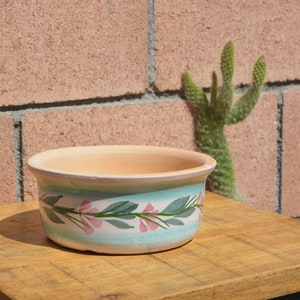 Puede incluir: Una maceta de terracota con un diseño floral azul claro y verde. La maceta está sobre una superficie de madera.