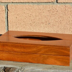 May include: A rectangular wooden tissue box with a smooth, polished finish. The box is a warm, reddish-brown colour and features a long, oval opening on the top for tissue dispensing. The box is sitting on a wooden surface.