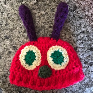 Peut inclure: Un bonnet crocheté rouge avec deux antennes violettes dressées, deux grands yeux circulaires aux anneaux extérieurs crème et aux centres vert foncé, ainsi qu'un petit nez circulaire vert foncé centré sous les yeux sur le devant.