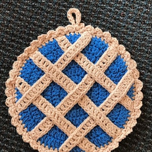 May include: A round, crocheted potholder featuring a blue center with a beige lattice crust design. The edges are finished with a scalloped beige border and a small loop at the top for hanging. The texture is consistent throughout the piece.