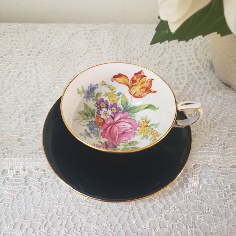 May include: A vintage teacup and saucer set. The teacup features a floral design with a pink rose and yellow, purple, and blue flowers. The cup and saucer are trimmed with gold. The saucer is black.