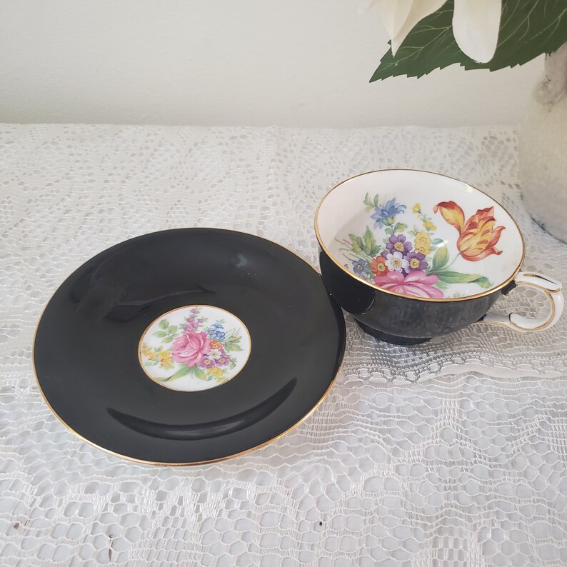May include: Black teacup and saucer set with gold trim. The teacup and saucer feature a floral design with pink, yellow, and blue flowers. The saucer has a central floral design, and the teacup has a matching interior floral pattern.