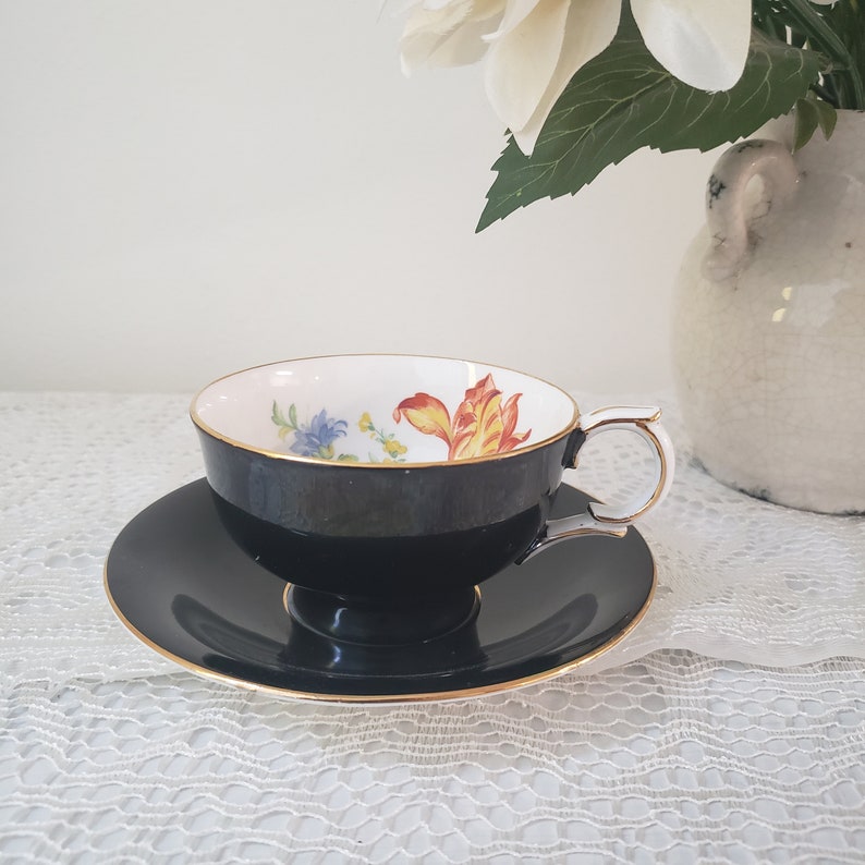May include: A black teacup and saucer set with a gold trim. The inside of the teacup features a floral design with yellow, orange, and blue flowers. The handle is white with a gold accent. The set is placed on a white lace tablecloth.
