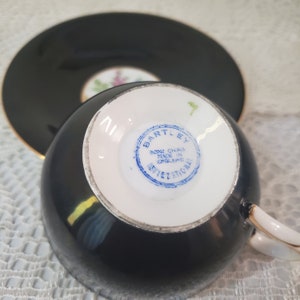 May include: Black teacup and saucer set with gold trim. The teacup has a white interior and a handle. The saucer is round. The bottom of the teacup has a blue stamp that reads "BARTLEY BONE CHINA MADE IN ENGLAND INTERNATIONAL".