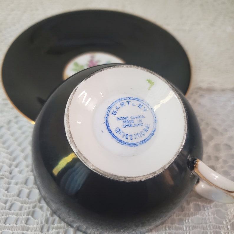 May include: Black teacup and saucer set with gold trim. The teacup has a white interior and a handle. The saucer is round. The bottom of the teacup has a blue stamp that reads "BARTLEY BONE CHINA MADE IN ENGLAND INTERNATIONAL".