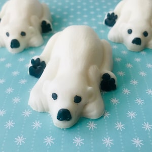 May include: Three white polar bear shaped soaps on a blue background with a snowflake pattern. The soaps are lying on their bellies with their paws visible.