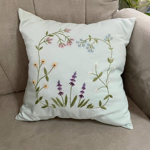 May include: A square light blue throw pillow featuring delicate hand-embroidered floral motifs. The design includes stems with small pink, light blue, yellow, and white flowers, along with three central stalks of purple lavender-like blossoms.