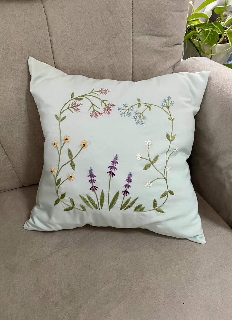 May include: A square light blue throw pillow featuring delicate hand-embroidered floral motifs. The design includes stems with small pink, light blue, yellow, and white flowers, along with three central stalks of purple lavender-like blossoms.