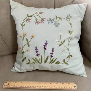 May include: A light blue square throw pillow featuring embroidered wildflowers in pink, blue, yellow, purple, and white. A wooden ruler placed at the base shows the pillow measures approximately 12 inches in width across the bottom edge.