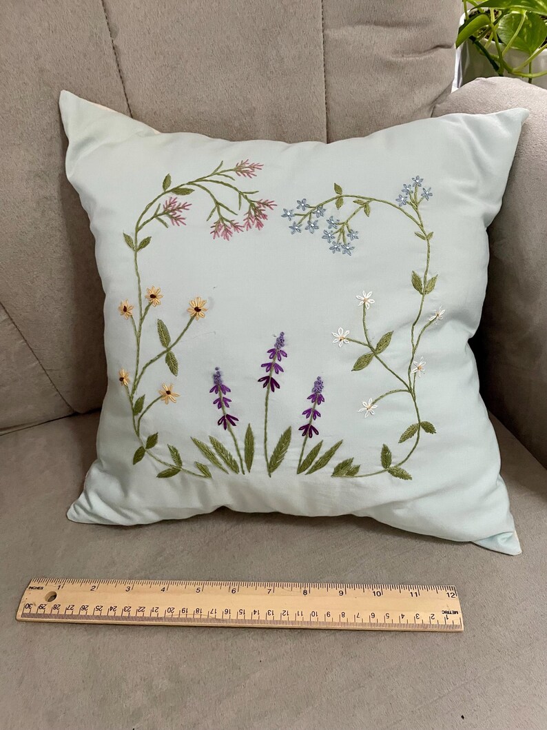 May include: A light blue square throw pillow featuring embroidered wildflowers in pink, blue, yellow, purple, and white. A wooden ruler placed at the base shows the pillow measures approximately 12 inches in width across the bottom edge.