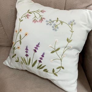 May include: A square light blue throw pillow featuring delicate hand-embroidered floral motifs. The design includes stems with small pink, light blue, yellow, purple, and white flowers arranged in a curved pattern across the front fabric surface.