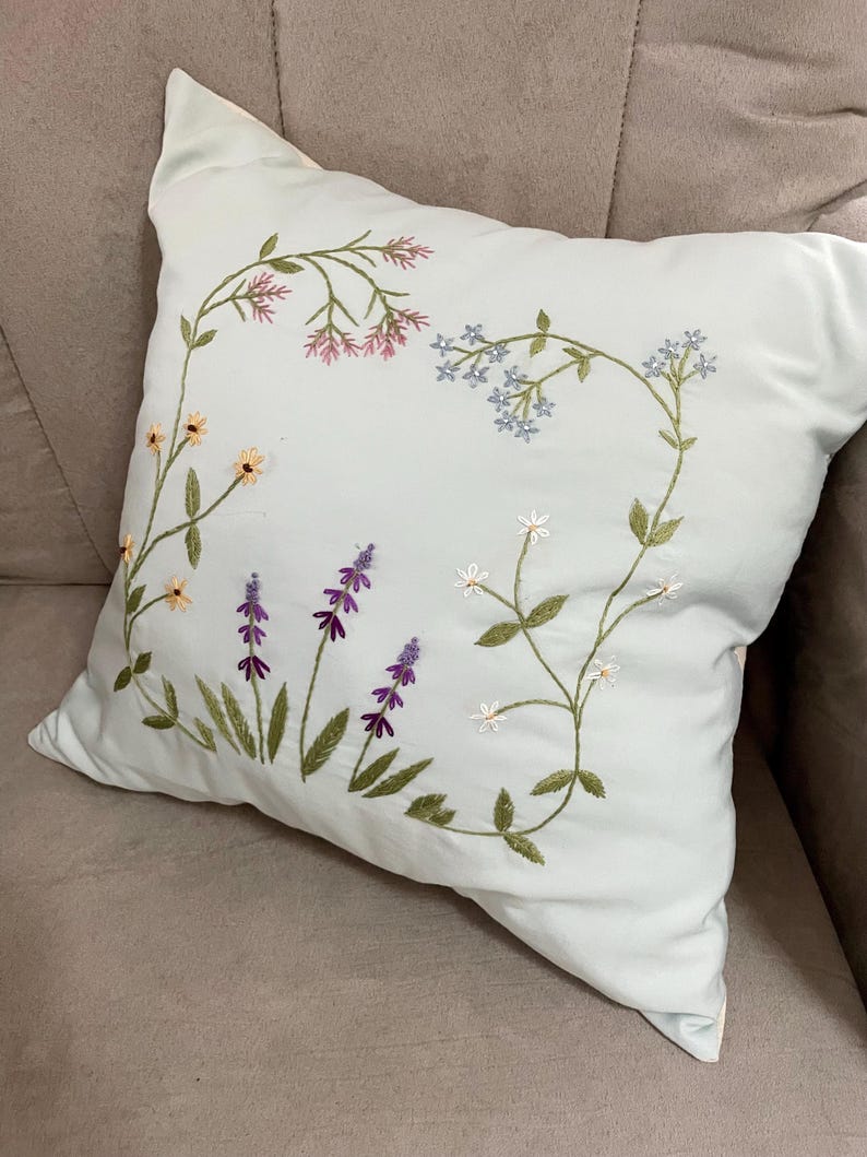 May include: A square light blue throw pillow featuring delicate hand-embroidered floral motifs. The design includes stems with small pink, light blue, yellow, purple, and white flowers arranged in a curved pattern across the front fabric surface.