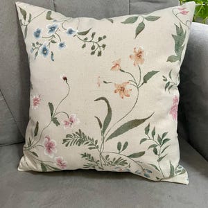 May include: A square throw pillow featuring a light beige fabric base with an embroidered botanical pattern. The design includes delicate flowers in shades of blue, peach, and pink, accented by green leaves and thin stems across the surface.