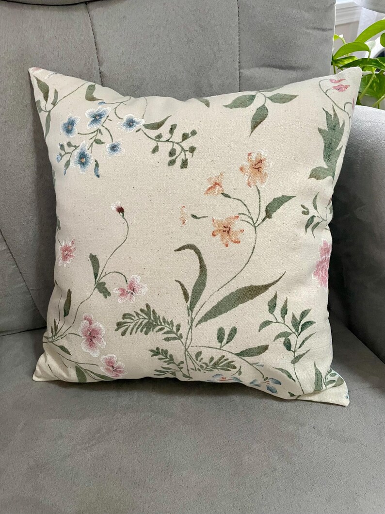 May include: A square throw pillow featuring a light beige fabric base with an embroidered botanical pattern. The design includes delicate flowers in shades of blue, peach, and pink, accented by green leaves and thin stems across the surface.