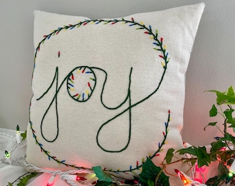 May include: A square off-white throw pillow featuring dark green embroidered cursive text that reads joy. The letter o is replaced by a circular wreath of colorful embroidered lights. A larger green embroidered wreath encircles the text.