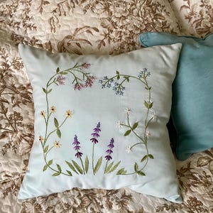 May include: A square light blue throw pillow featuring delicate hand-embroidered wildflowers in shades of purple, yellow, white, and blue. The floral design is arranged in a rectangular frame pattern with green stems and leaves on the fabric.