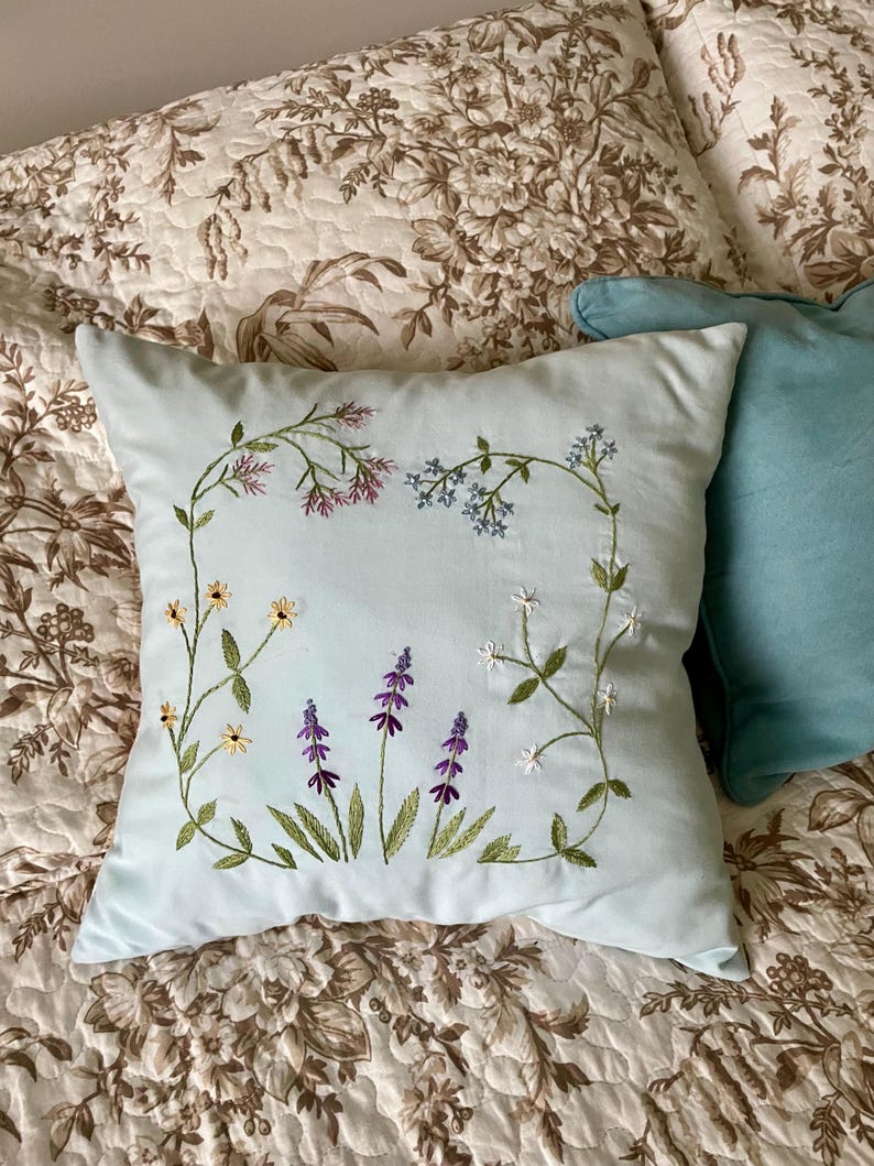 May include: A square light blue throw pillow featuring delicate hand-embroidered wildflowers in shades of purple, yellow, white, and blue. The floral design is arranged in a rectangular frame pattern with green stems and leaves on the fabric.