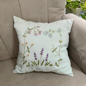 May include: A square light blue throw pillow featuring a central hand-embroidered design of various wildflowers. The embroidery includes delicate stems with pink, blue, yellow, white, and purple flowers arranged in a rectangular floral frame pattern.