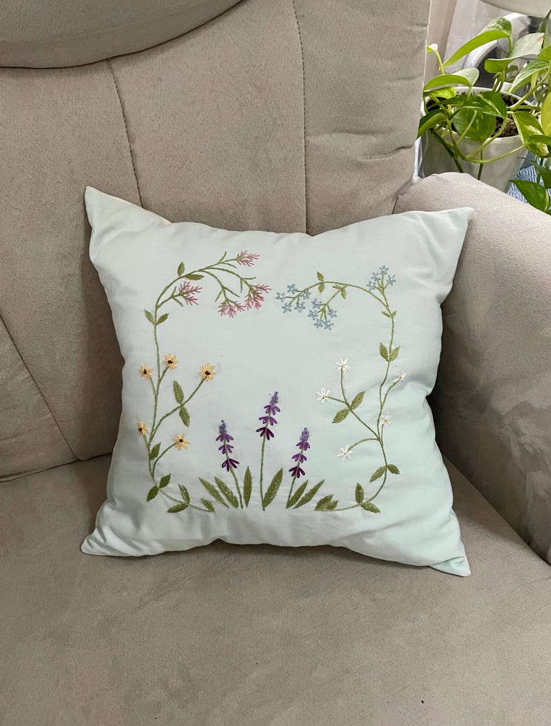 May include: A square light blue throw pillow featuring a central hand-embroidered design of various wildflowers. The embroidery includes delicate stems with pink, blue, yellow, white, and purple flowers arranged in a rectangular floral frame pattern.