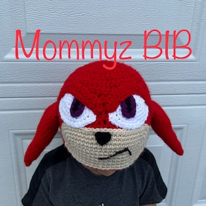 May include: A red and white crocheted hat with a cartoon face. The hat has purple eyes, a black nose, and a white mouth. The hat is worn by a child wearing a grey shirt with the word "MAN" on it. The text "Mommyz BIB" is written in red at the top of the image.