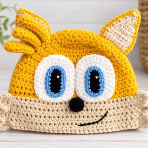 May include: A yellow and beige crochet hat featuring a character face with large white eyes, blue irises, black pupils, a small black nose, and a stitched black smile. The hat includes two pointed ears and a tuft of yellow hair on the top left side.
