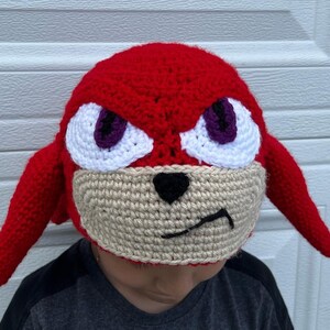 May include: A red crocheted hat with a white face and purple eyes. The hat has two long red ears.