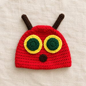 Peut inclure: Un bonnet crocheté rouge avec deux antennes marron sur le dessus, deux grands yeux circulaires aux centres vert foncé bordés de jaune vif, et un petit nez circulaire marron foncé centré sous les yeux sur le devant.