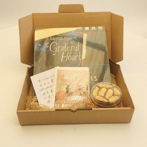 Pode incluir: Uma caixa de presente castanha contendo um livro intitulado "Kareem Khaleel and The Grateful Heart", um cartão com o texto "Happy Eid" e um frasco de biscoitos. A caixa também inclui um cartão com texto árabe.