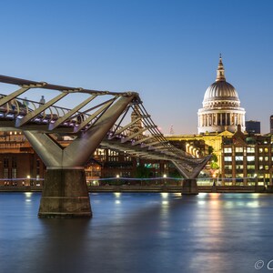 May include: A view of the Millennium Bridge in London at dusk, with St. Paul's Cathedral in the background. The bridge is lit up, and the water is reflecting the lights.