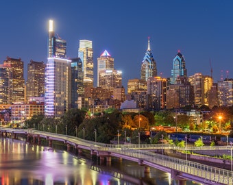 May include: A panoramic view of the Philadelphia skyline at night, with the city's iconic skyscrapers illuminated against a dark blue sky. The buildings are reflected in the water of the Delaware River, creating a beautiful and symmetrical scene.