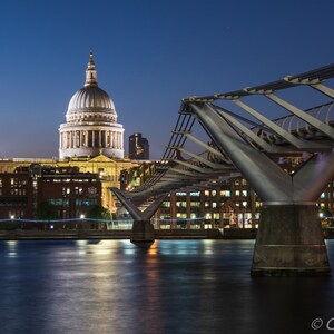 May include: Nighttime view of London's skyline featuring St. Paul's Cathedral and the Millennium Bridge. The bridge's steel structure contrasts with the cathedral's dome. The dark blue sky and the water of the Thames River reflect the city lights.