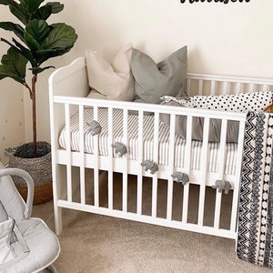 May include: A white crib with gray and white striped bedding, elephant-shaped decorations, and a black and white patterned blanket. The wall features framed animal prints and a black name sign that reads "Harrison". A gray baby bouncer and a potted plant are also in the room.