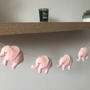 May include: A garland of pink felt elephants strung on a white and gray twine. Each elephant has closed eyes and is facing to the right. The garland is hanging in front of a wooden shelf with a candle and a potted succulent.