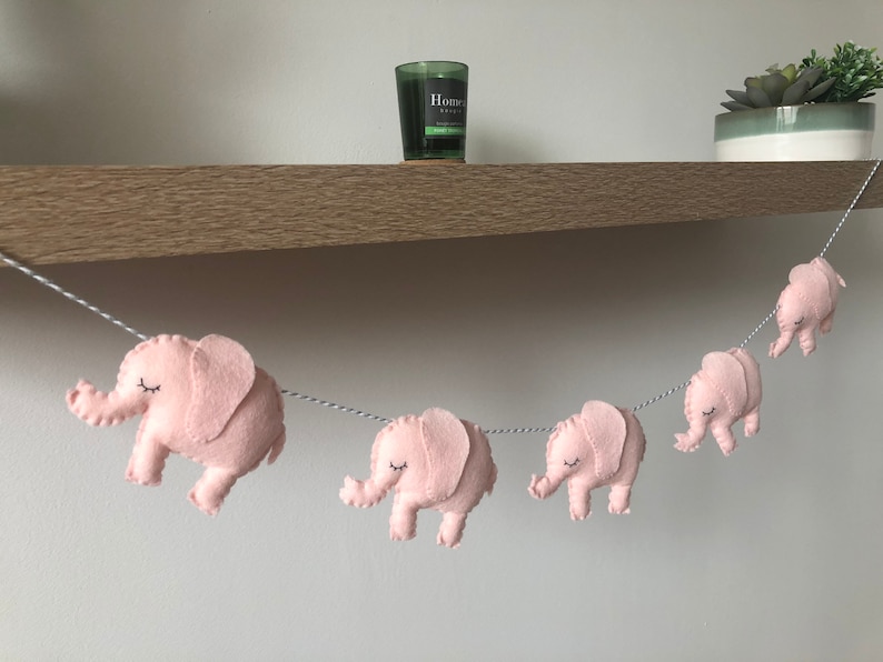 May include: A garland of pink felt elephants strung on a white and gray twine. Each elephant has closed eyes and is facing to the right. The garland is hanging in front of a wooden shelf with a candle and a potted succulent.