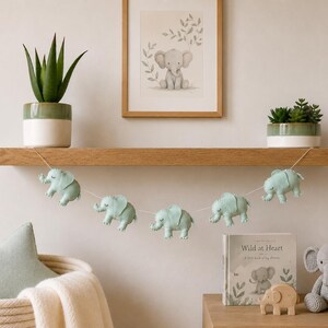 May include: A nursery shelf displays a garland of light blue elephant ornaments, a framed elephant print, and a stuffed elephant toy. A potted succulent and a book titled "Wild at Heart" are also visible.