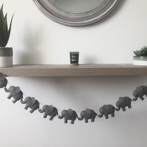 May include: A gray felt elephant garland strung on a white string, hanging below a wooden shelf. The garland is displayed with two potted plants and a small candle. The garland is a decorative item.
